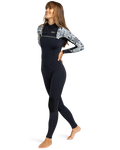 A woman smiles as she walks forward barefoot on a plain white background, wearing the Roxy 3/2mm Swell Chest Zip Fullsuit in black with white patterned sleeves. One arm is bent behind her back.