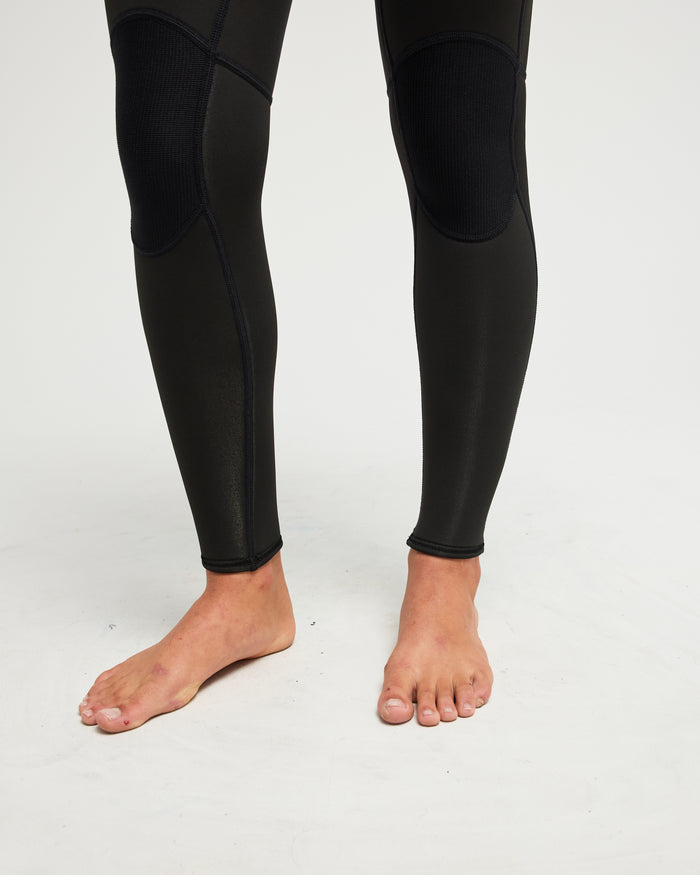 A person stands barefoot on a white surface, wearing the Roxy 3/mm Prologue Back Zip (Flatlock) Wetsuit in black, which fits snugly and covers the legs up to the ankles.