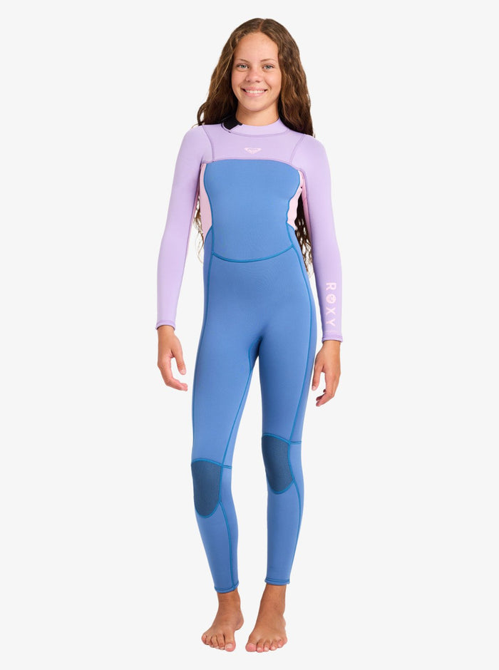 A young girl stands barefoot and smiling against a plain white background, wearing the Roxy 4/3mm Prologue Girls’ Back Zip Wetsuit featuring long blue and lavender sleeves, knee pads, and a Roxy logo on the arm.