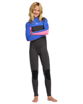 A smiling young girl with long blonde hair stands barefoot in the Roxy 4/3mm Prologue Girls' Back Zip Wetsuit, made from stretch neoprene and recycled polyester with blue, pink, and black panels, posing confidently against a white background.