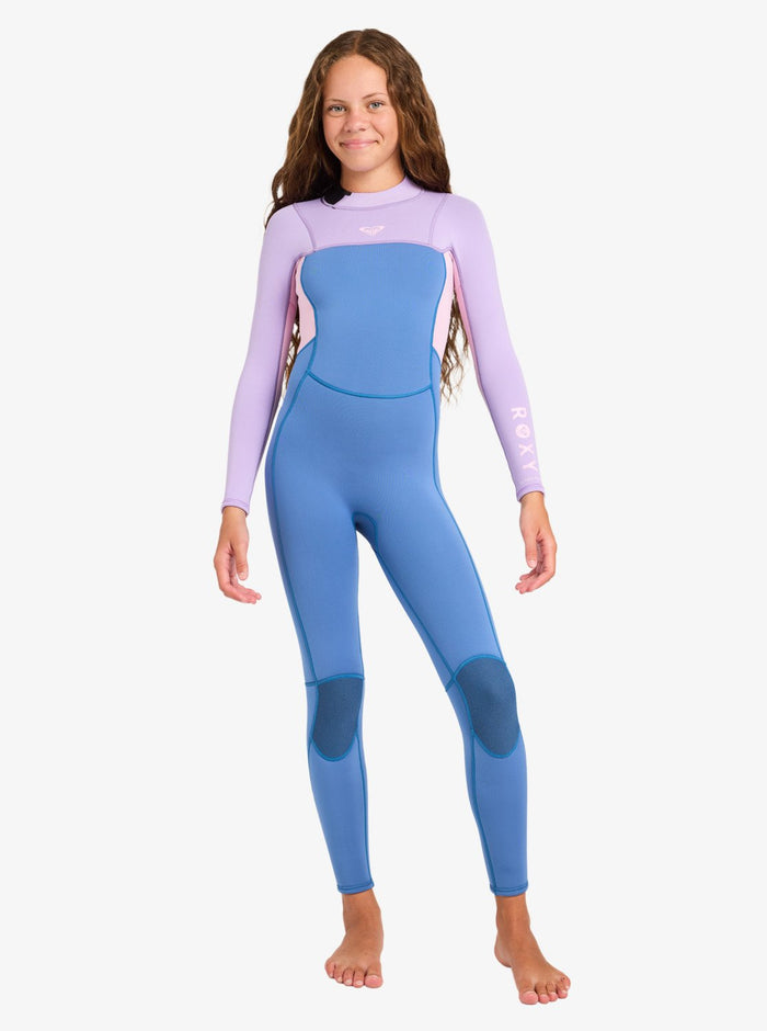 A young girl with long brown hair stands barefoot on a white background, smiling softly as she models the Roxy 4/3mm Prologue Girls’ Back Zip Wetsuit in shades of blue and lavender.