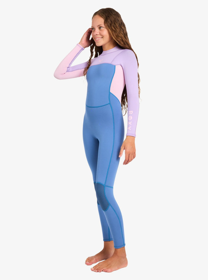 A smiling woman stands barefoot in the Roxy 4/3mm Prologue Girls’ Back Zip Wetsuit, which features long sleeves in blue and light purple. She has long, wavy hair and touches her head with one hand, set against a plain white background.