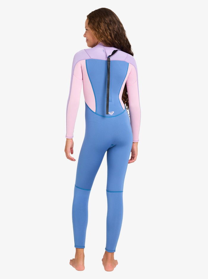 A girl with long brown hair stands barefoot and facing away from the camera, wearing the Roxy 4/3mm Prologue Girls’ Back Zip Wetsuit in blue and pink, featuring a black zipper running down the back.