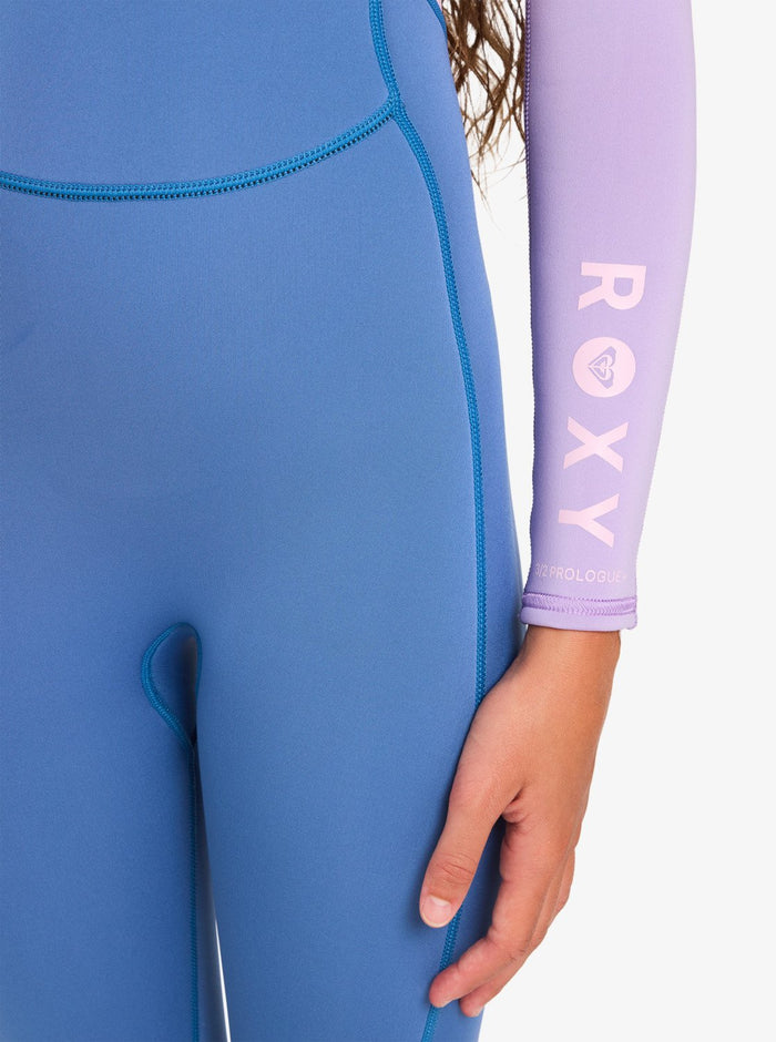 A person wearing the Roxy 4/3mm Prologue Girls’ Back Zip Wetsuit, featuring a blue body and a lavender sleeve with “ROXY” printed in white. Only the lower torso, right arm, and hand are visible.