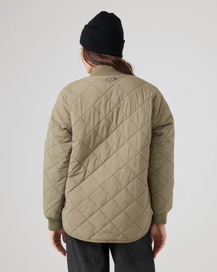 A person stands facing away, wearing the Roxy Aurora Quilted Bomber in khaki, paired with a black beanie and dark pants, highlighting the jacket’s small embroidered detail at the neck on the back.