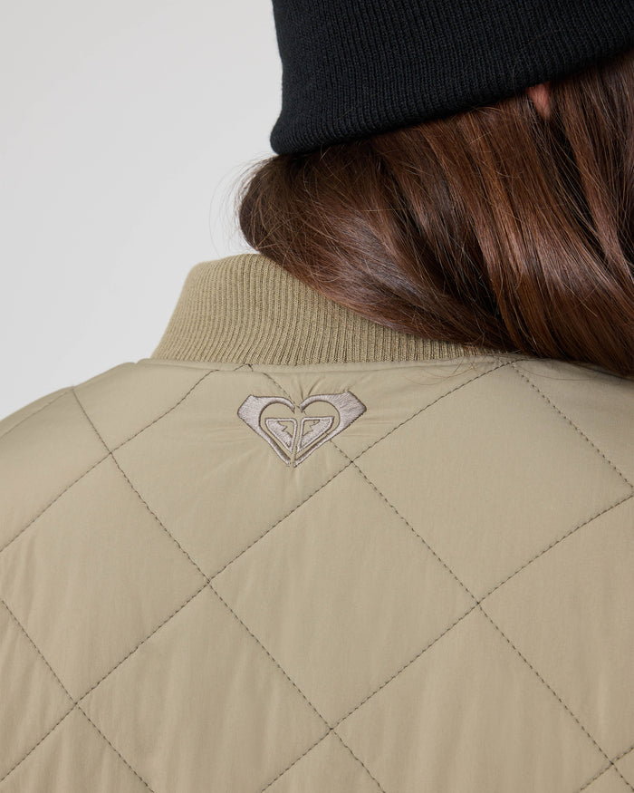 Close-up of a person with long brown hair wearing a black knit beanie and the Roxy Aurora Quilted Bomber in beige, featuring a small embroidered heart-shaped logo on the upper back.