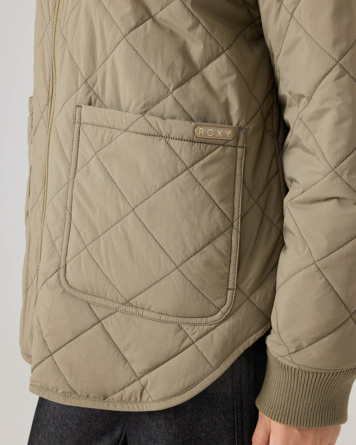 Close-up of the Aurora Quilted Bomber by Roxy in khaki, featuring a large front pocket and a small Roxy label. The jacket is styled with dark pants, with only the torso and part of the arm visible.