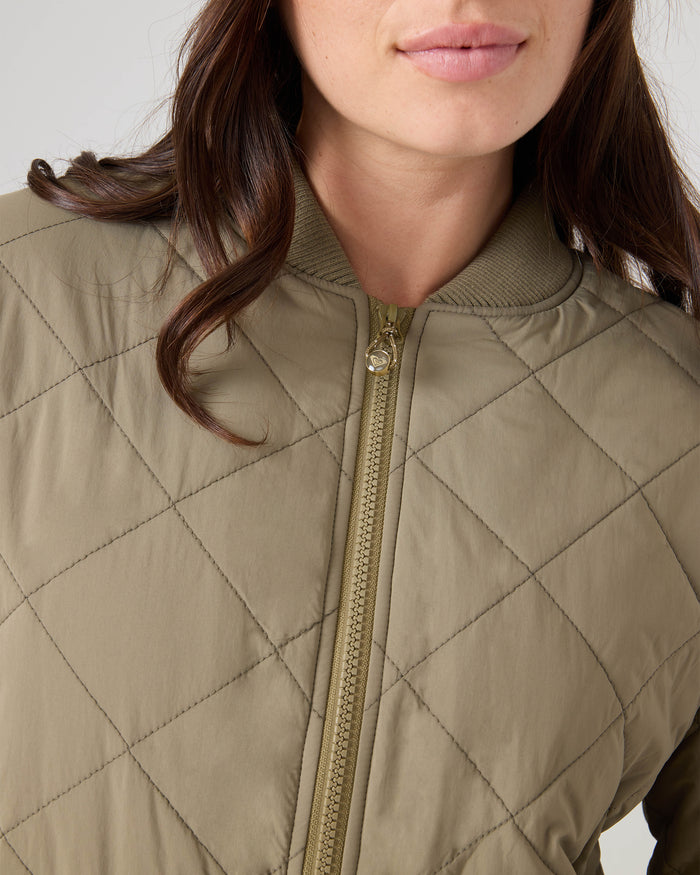 A woman is shown from the shoulders up wearing the Roxy Aurora Quilted Bomber in light olive green, featuring a gold zipper. Her brown wavy hair frames her face, and she has a neutral expression.