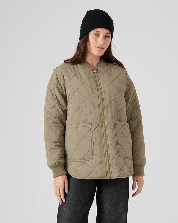 A person is wearing the Roxy Aurora Quilted Bomber, a stylish olive jacket with front pockets, paired with a black beanie and dark pants, standing against a plain light background.