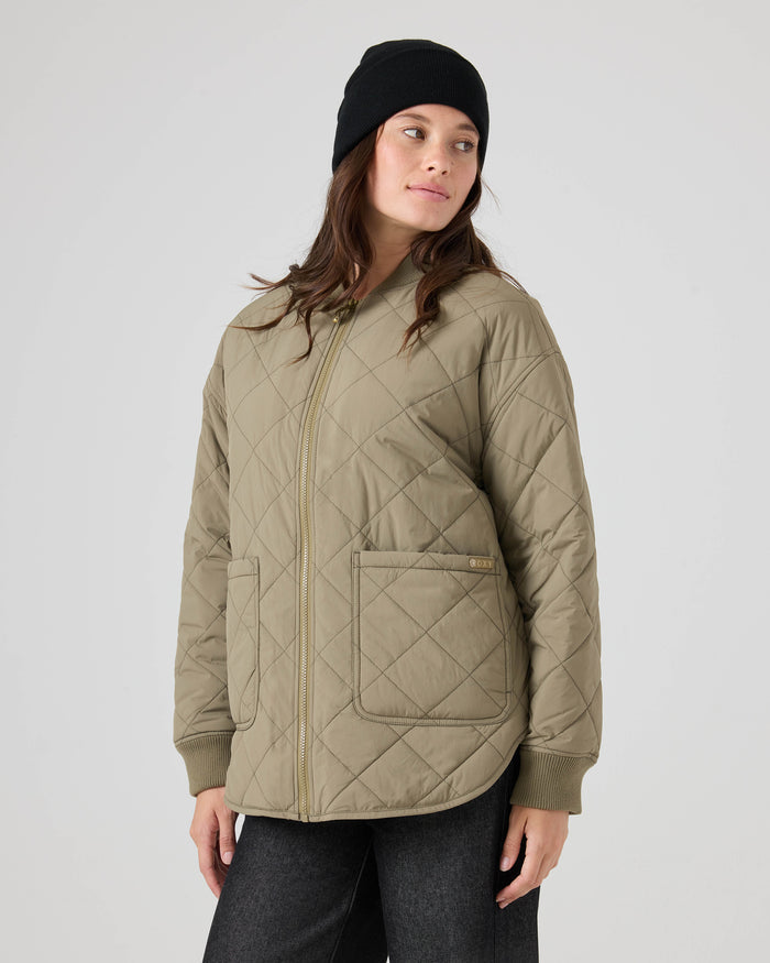 A woman wearing a black beanie and the Roxy Aurora Quilted Bomber in light olive, featuring front pockets, stands against a plain white background, looking to her left.