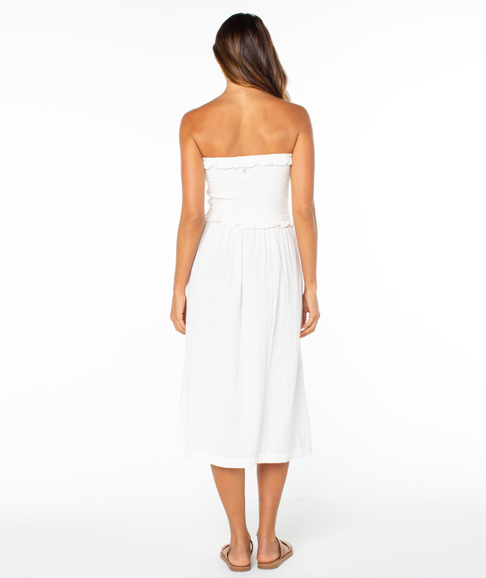 A woman with long hair stands facing away, wearing the Roxy Avalon Tube Midi Dress styled with sandals and a breezy skirt, set against a plain white background.