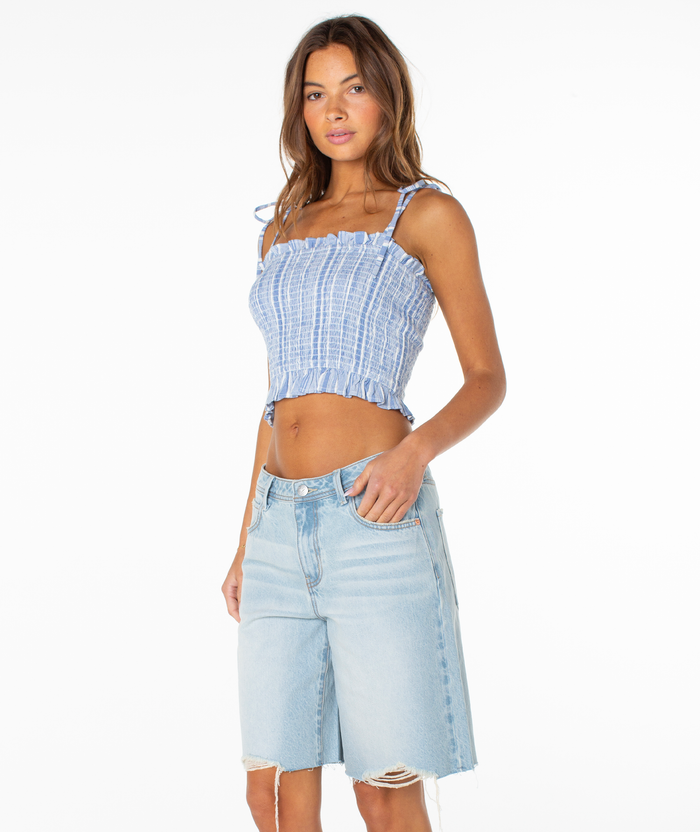 A woman with long brown hair and a neutral expression wears the Roxy Avalon Tube Top, featuring blue and white stripes and tied straps, paired with light blue knee-length distressed denim shorts. She faces slightly left against a white background.