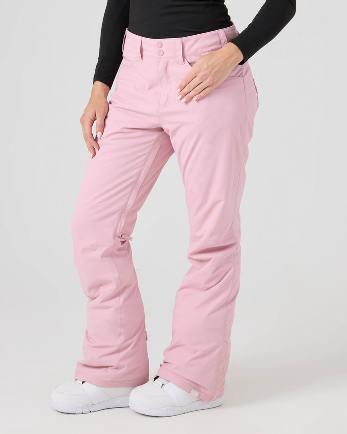 A person wearing Roxy Backyard Pants in Zephyr—a light pink color—made from recycled polyester, paired with a black long-sleeve top and white snow boots, stands against a plain light background. Only the lower torso and legs are visible.