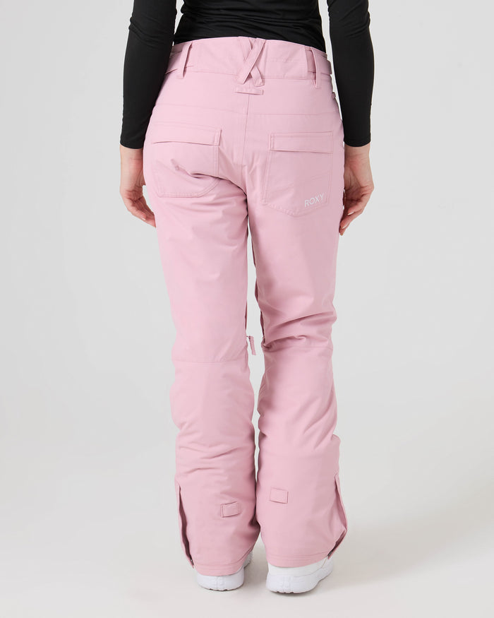 A person is wearing Roxy Backyard Pants in light pink—women’s waterproof ski pants crafted from recycled polyester—paired with a black long-sleeve top and white shoes. The image shows the back view, featuring visible pockets and a subtle Roxy logo on the right pocket. The background is plain and light-colored.