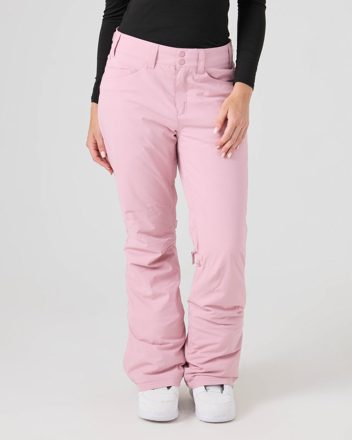 A person wearing Roxy Backyard Pants in light pink made from recycled polyester, paired with a black long-sleeve top and white sneakers, standing against a plain light background.