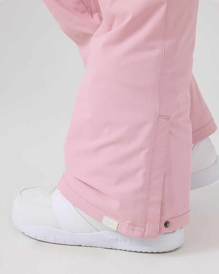 A person wearing Roxy Backyard Pants in light pink, made from recycled polyester and featuring a snap button detail near the ankle, paired with white sneakers and standing on a light surface.