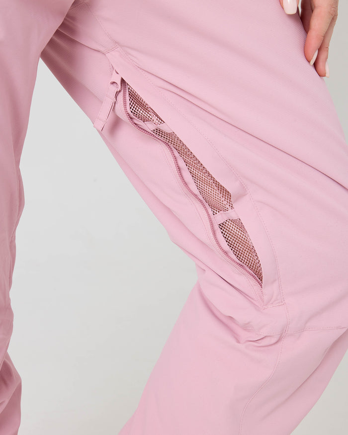 Close-up of a person wearing the Roxy Backyard Pants in light pink, featuring a partially open zippered mesh vent that reveals the inner mesh lining. The person’s manicured hand rests near the waistband of these stylish waterproof ski pants.