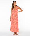 A woman with long wavy hair poses confidently in a Roxy Beach Day Maxi Dress, featuring summery pink and orange florals, a deep V-neck, and brown sandals against a white background.