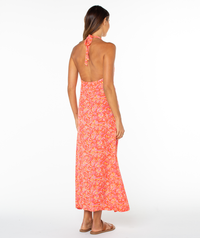 A woman faces away in the Roxy Beach Day Maxi Dress, featuring a halter neckline and bright pink and orange floral print. She has long hair, wears flat sandals, and stands against a plain white background.