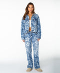 A woman stands in a studio against a white background, wearing the Roxy Big Tide Jacket in Flora Fade Print Denim Wash with matching jeans, styled with a white crop top and beige shoes.