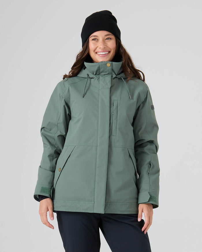 A smiling person wears the Roxy Billie Jacket in Duck Green, paired with black pants and a black beanie, standing against a plain light background.