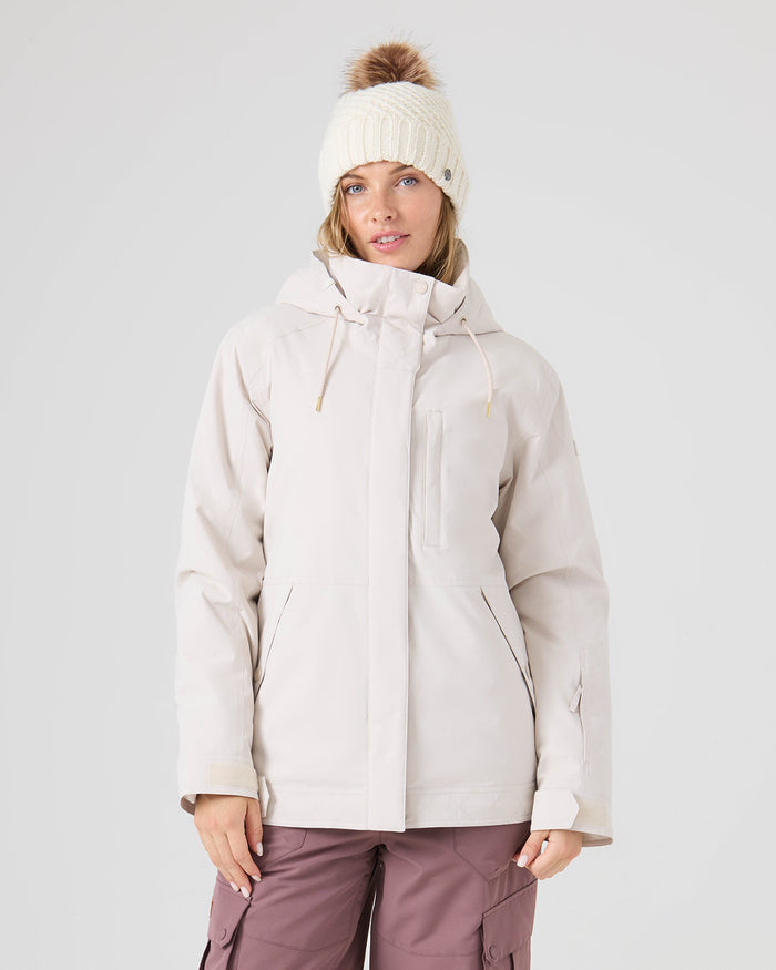 A woman wearing a cream beanie with a brown pom-pom, a light beige Roxy Billie Jacket in Moonbeam with microripstop protection, and mauve pants stands facing forward against a plain light gray background.