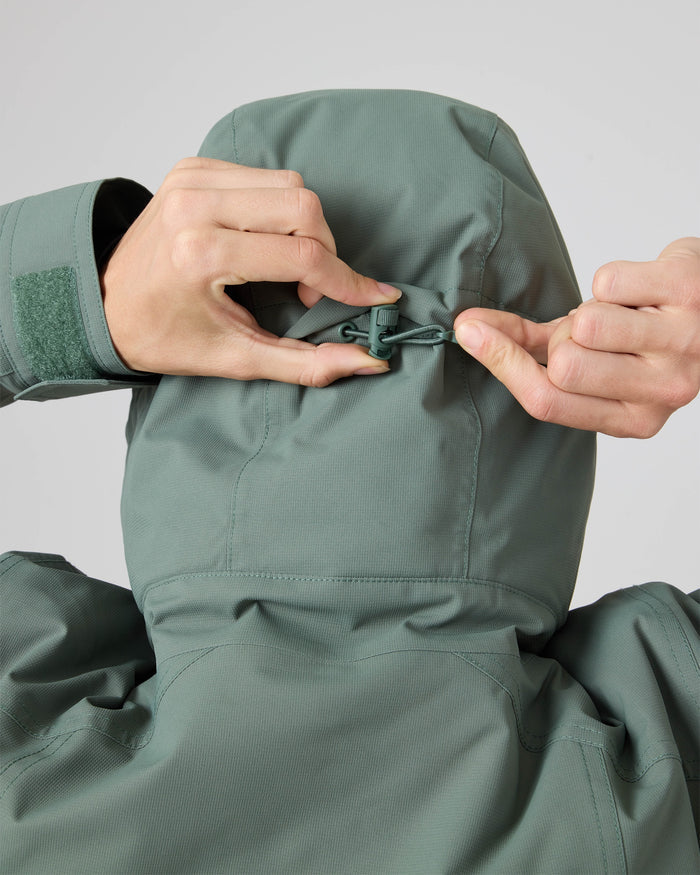 A person adjusts the hood of a green Roxy Billie Jacket using a drawstring at the back, with both hands visible and the face not shown. Crafted from recycled polyester with durable 10K/5K microripstop fabric, the women’s snowboard jacket combines style and performance for winter adventures.