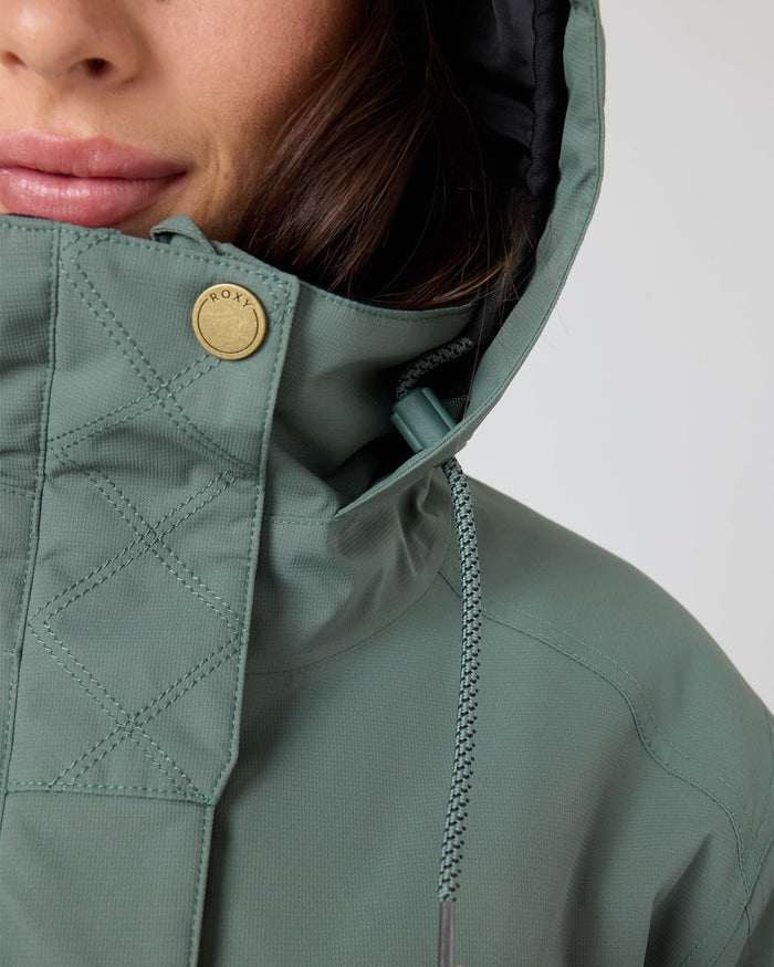 Close-up of a person wearing the Billie Jacket by Roxy—a women’s snowboard jacket in green, made from recycled polyester. The jacket features a braided drawstring and a gold button engraved with ROXY. Only the lower half of the face is visible.