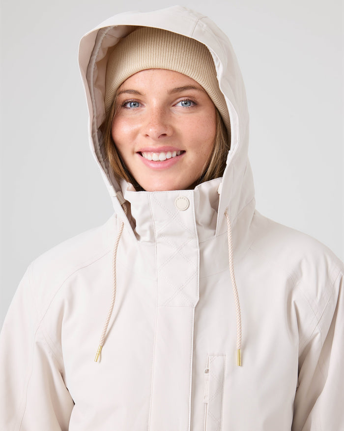 A person wearing the Roxy Billie Jacket in beige with microripstop protection and a tan beanie smiles at the camera against a light gray background.