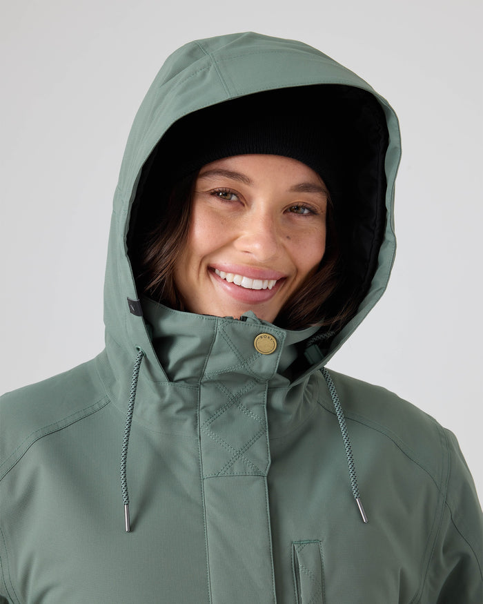 A person smiling, wearing the Roxy Billie Jacket in sage green—crafted from 10K/5K microripstop recycled polyester—over a black beanie, with the hood up, set against a plain light background.