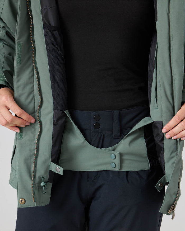 A person wearing the Roxy Billie Jacket, a green women’s snowboard jacket crafted from recycled polyester with 10K/5K microripstop fabric, over a black shirt displays the jacket’s interior snow skirt and snaps, while dark pants are visible below.