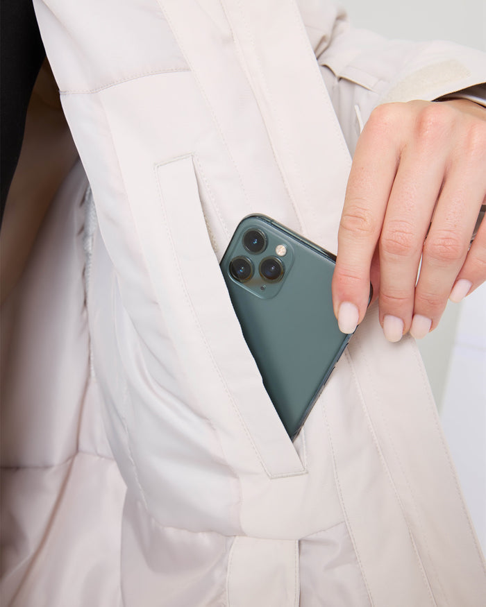 A person with manicured nails is placing a smartphone with three camera lenses into the pocket of a light-colored Roxy Billie Jacket, highlighting both style and practicality with its microripstop protection.
