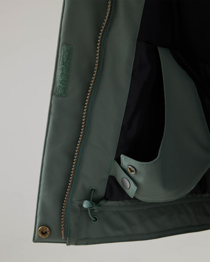 Close-up of the bottom edge of the Roxy Billie Jacket in green, highlighting its gold zipper, metal snap button, drawstring, and part of the interior black lining crafted from recycled polyester 10K/5K microripstop.