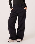 A person stands against a plain light background with hands in pockets, wearing the Bleeker Pants by Roxy—loose-fitting black pants inspired by women’s snowboard styles. The pants feature an elastic waistband, belt, and visible zippers for style and function. Crafted from waterproof-breathable materials with PFAS-free finishes, they provide both comfort and sustainability. The look is completed with a black long-sleeve top and white sneakers.