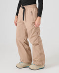 A person is wearing the Roxy Bleeker Pants in Chanerelle—a beige, loose-fit style made from recycled nylon—paired with a black belt, a black long-sleeve top, and beige shoes with blue details, standing against a plain light background.