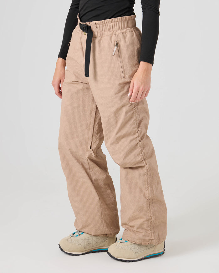 A person is wearing the Roxy Bleeker Pants in Chanerelle—a beige, loose-fit style made from recycled nylon—paired with a black belt, a black long-sleeve top, and beige shoes with blue details, standing against a plain light background.