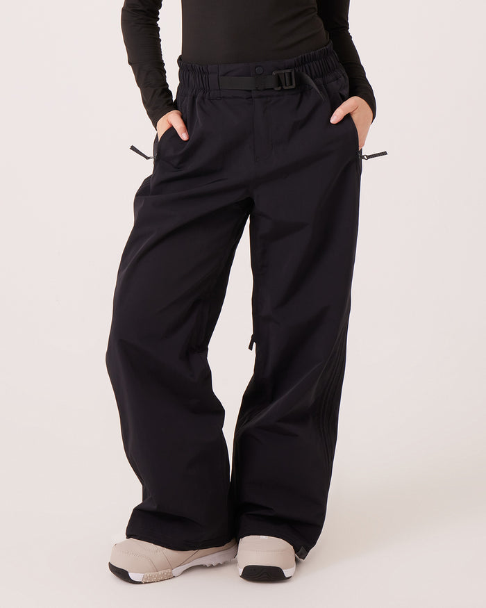 A person wearing the Roxy Bleeker Pants—loose-fitting, black, waterproof-breathable trousers with an elastic waistband, side pockets, and a belt—paired with a long-sleeve black top and white chunky sneakers. Only the lower half of the body is visible.