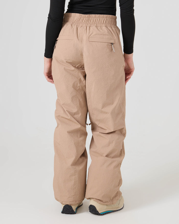 A person stands with their back to the camera on a plain light background, wearing Roxy’s Bleeker Pants—beige, loose-fit recycled nylon pants featuring zippered back pockets—paired with a black long-sleeve top and light-colored sneakers.