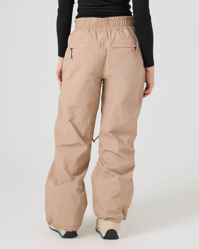 Model wears the Roxy Bleeker Pants in a loose-fit beige style featuring back pockets, paired with a black long-sleeve top and white sneakers; shown from the back against a plain light background.