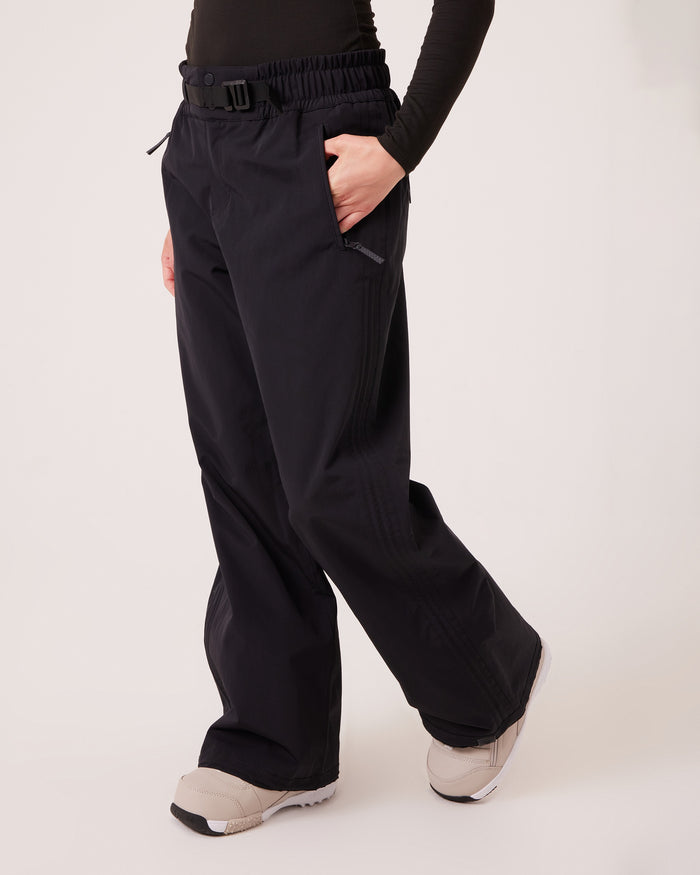 Wearing the Roxy Bleeker Pants—black, wide-leg, and crafted from PFAS-free fabric—a person stands with one hand in their pocket, paired with a black long-sleeve top and light-colored sneakers, facing slightly to the side.