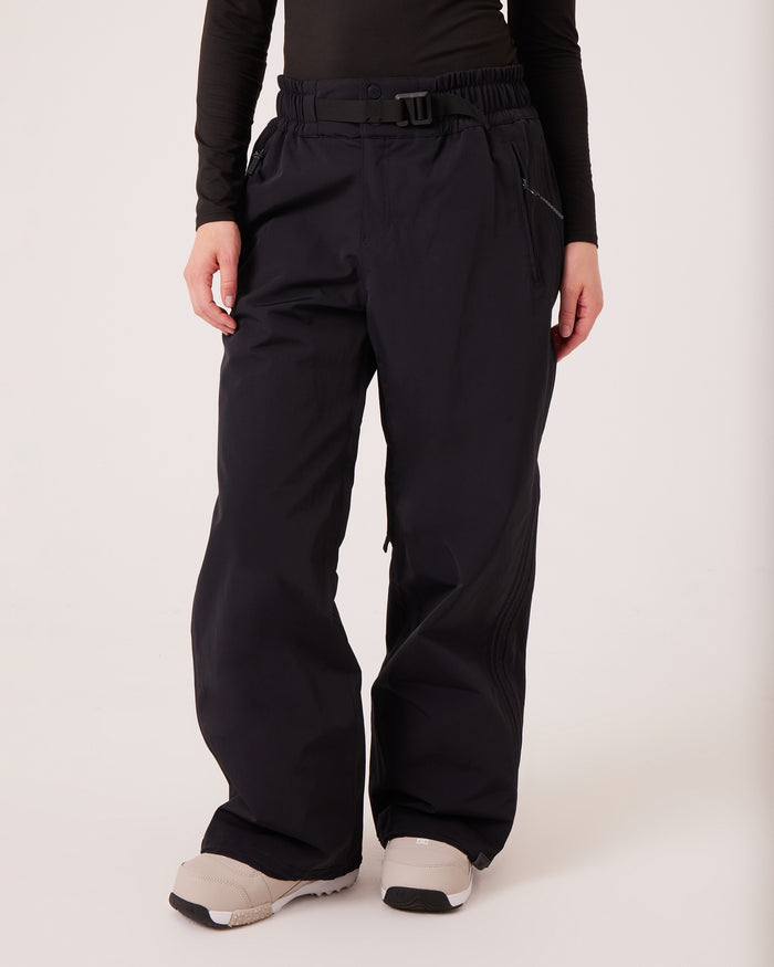 A person wearing loose-fitting, PFAS-free Roxy Bleeker Pants, a black long-sleeve top, and light beige winter boots, standing against a plain white background.