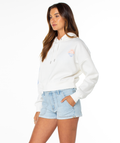 A woman stands against a white background wearing the Roxy Breakwater Hoodie, a relaxed cotton blend with a sun and heart graphic, paired with light blue denim shorts. Her hands are in her pockets and she looks slightly to the side.
