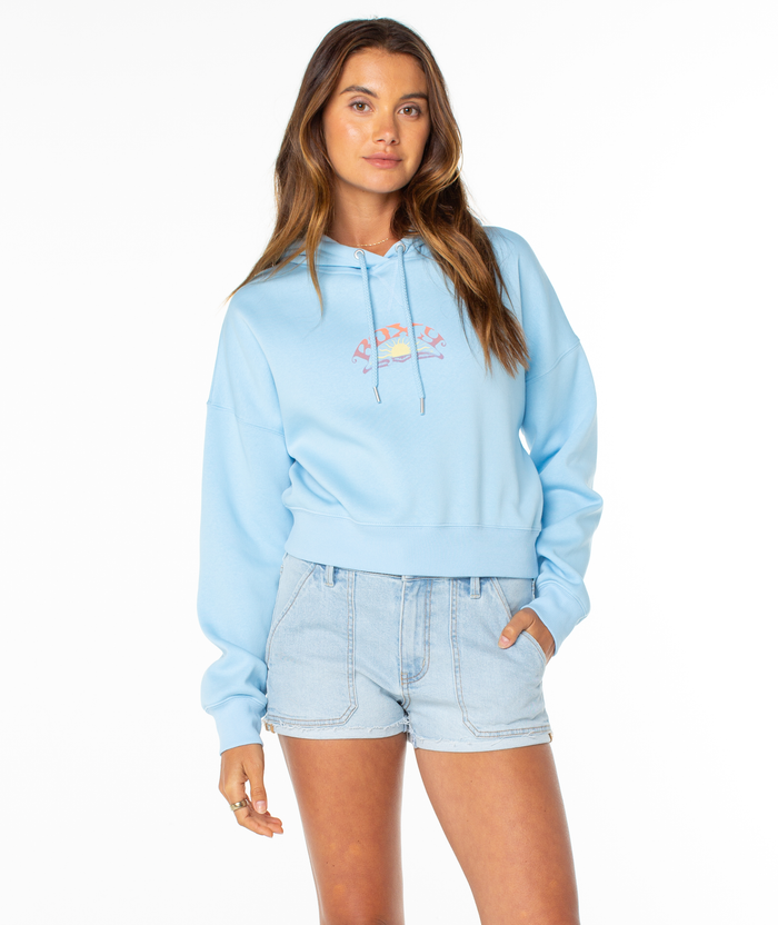A woman wears the Roxy Breakwater Hoodie, a relaxed cotton blend with Roots text and rainbow front graphic in a cropped fit, paired with light blue denim shorts, standing against a plain white background.