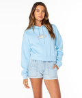 A woman wears the Roxy Breakwater Hoodie, a relaxed cotton blend with Roots text and rainbow front graphic in a cropped fit, paired with light blue denim shorts, standing against a plain white background.