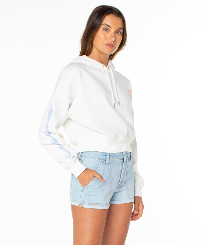 A woman stands against a white background wearing the Roxy Breakwater Hoodie, a relaxed cotton-blend with sleeve designs, and light denim shorts. She has one hand in her pocket and a neutral expression.