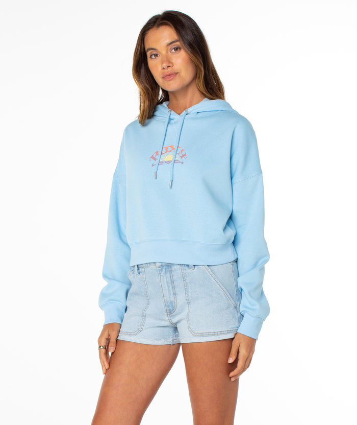 A woman models the Roxy Breakwater Hoodie in light blue, featuring a small colorful chest graphic, paired with light denim shorts. She stands against a white background and looks at the camera with a neutral expression.