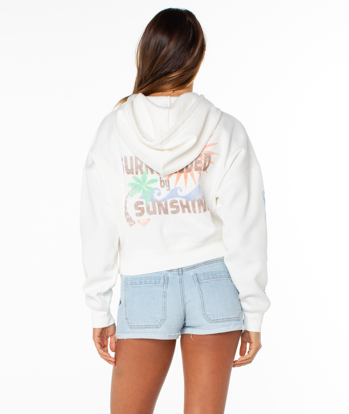 A woman with long brown hair wears the Roxy Breakwater Hoodie, a cropped style with a colorful beach graphic, paired with light blue denim shorts. She faces away from the camera against a white background.