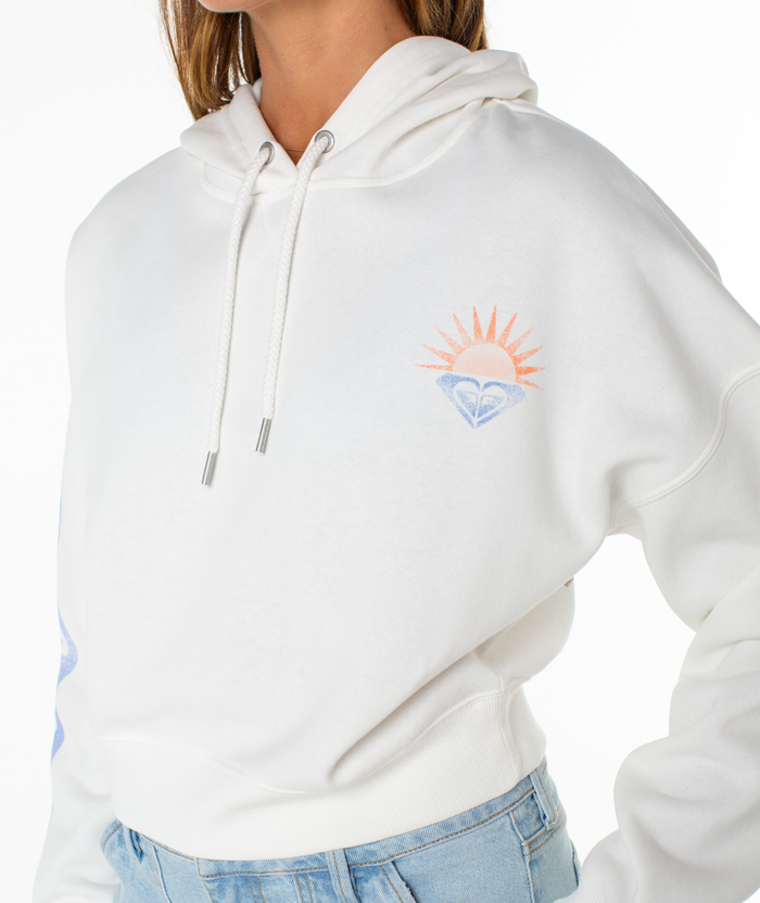 A person in a white Roxy Breakwater Hoodie with a drawstring, small orange sun and blue wave graphic on the chest, cropped relaxed cotton blend fit, styled with light blue jeans.