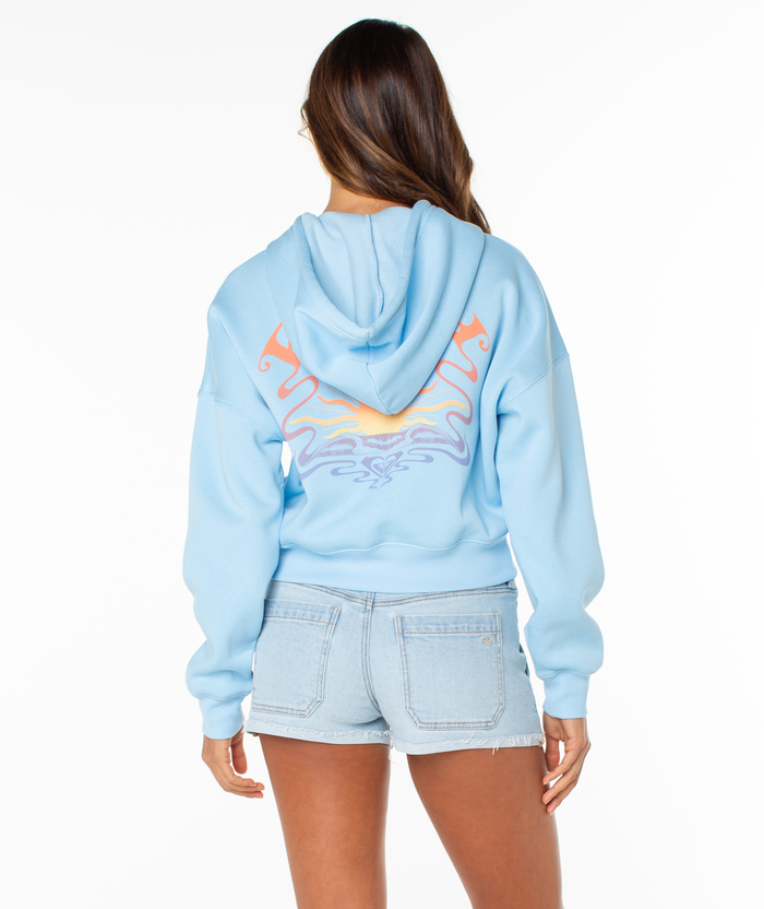 A woman faces away in a light blue Roxy Breakwater Hoodie with a colorful sun graphic on the back and light denim shorts. The relaxed cotton blend hoodie has a cropped fit, and her long brown hair falls against a white background.