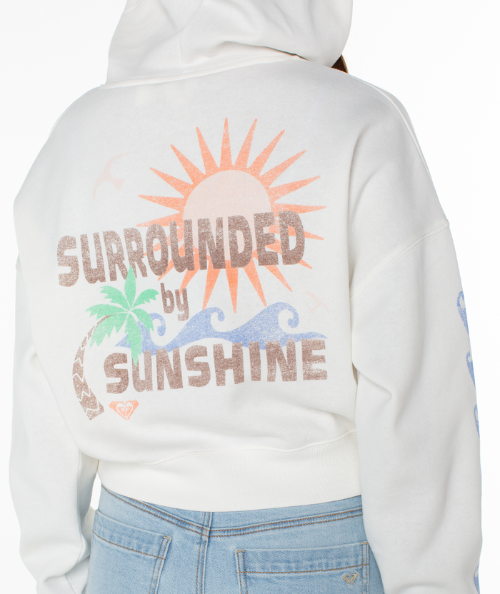 A person wears the relaxed cotton blend Roxy Breakwater Hoodie, featuring a back design with a sun, palm tree, waves, and "Surrounded by Sunshine" in pastel colors, paired with light blue jeans.